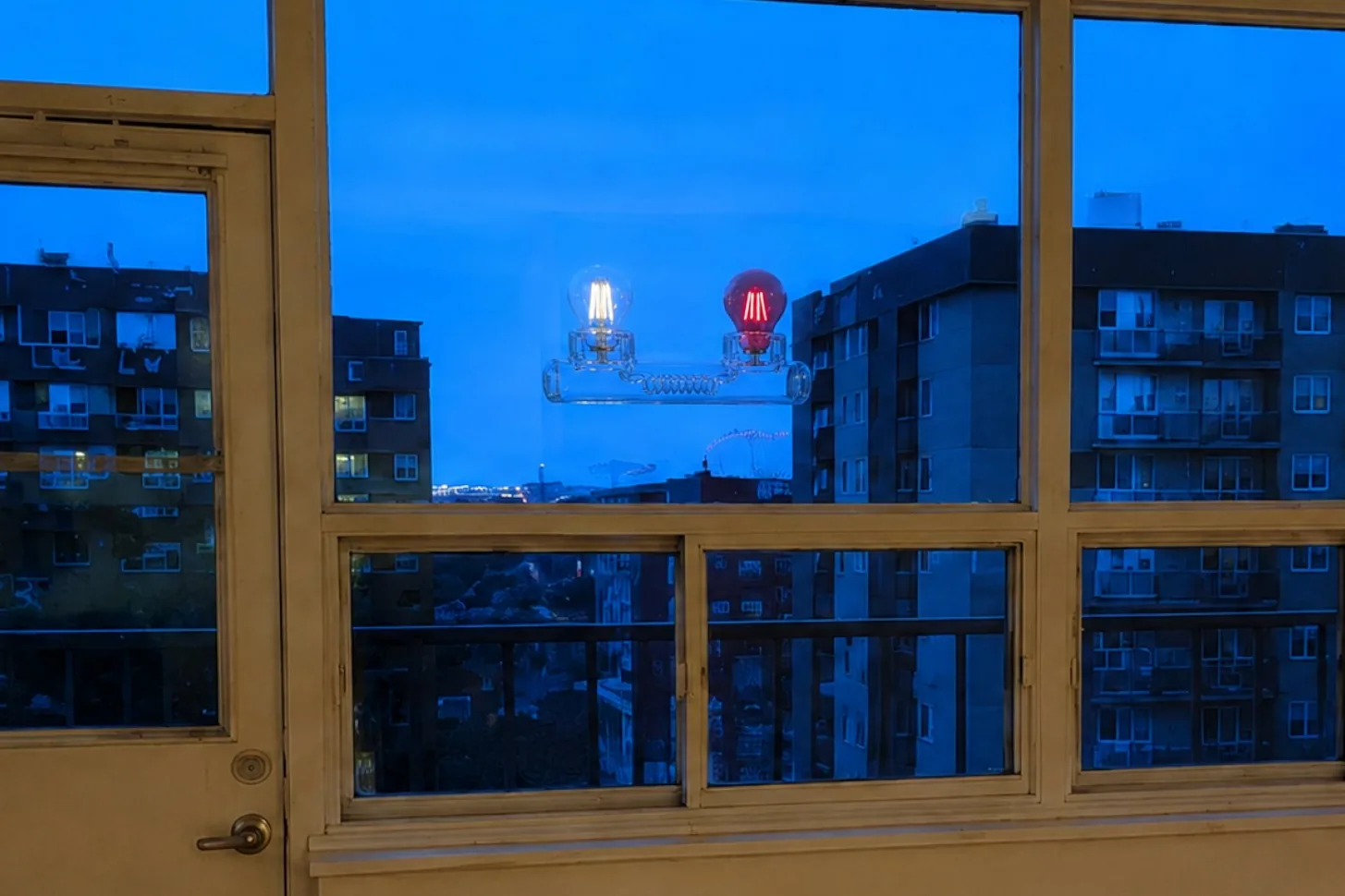 A recreation of a glass-tube object topped by two light bulbs, one clear and one red, hovering outside an apartment window at dusk over a Toronto street